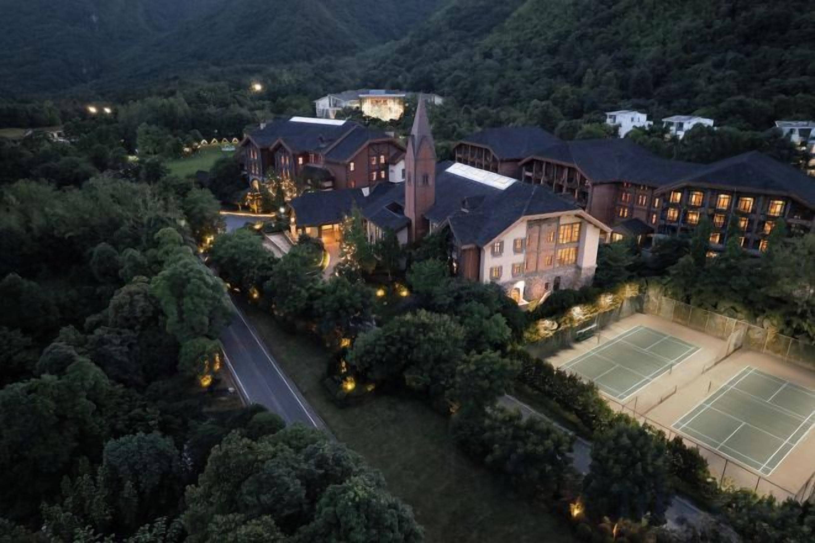 A large hotel with multiple buildings, surrounded by dense forest, featuring tennis courts and a well-lit entrance.