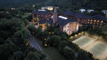 A large hotel with multiple buildings, surrounded by dense forest, featuring tennis courts and a well-lit entrance.