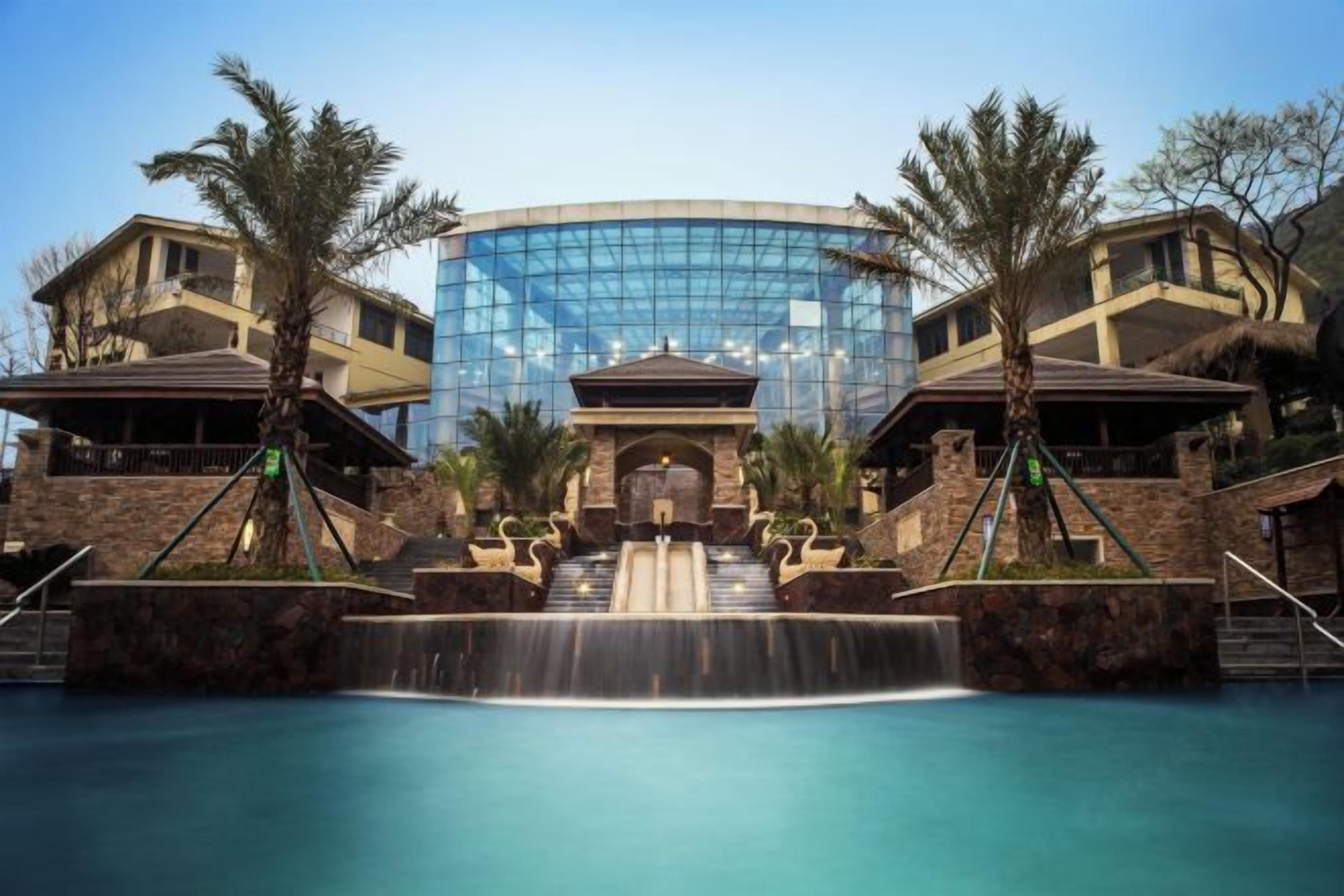 A hotel with a central glass building, a pool with a waterfall feature, and palm trees.
