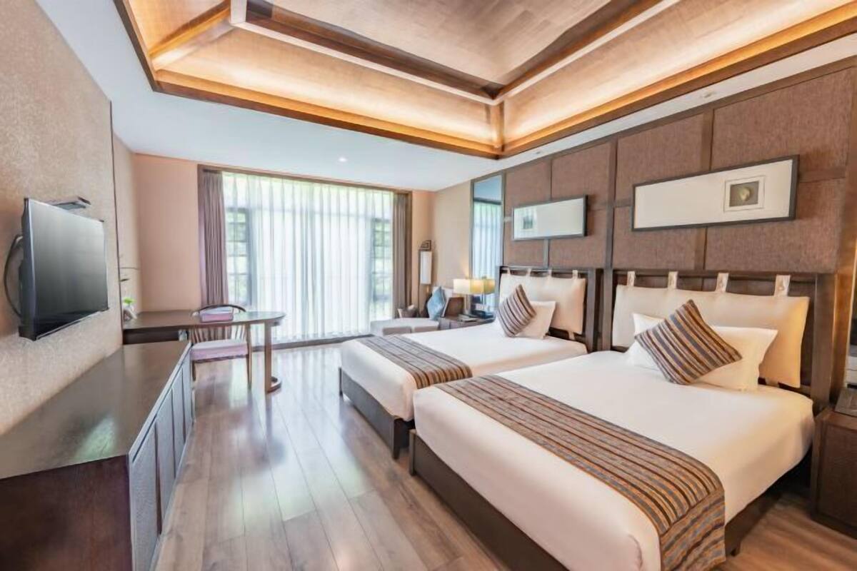 A hotel room with two beds, a TV, a desk, and a large window.