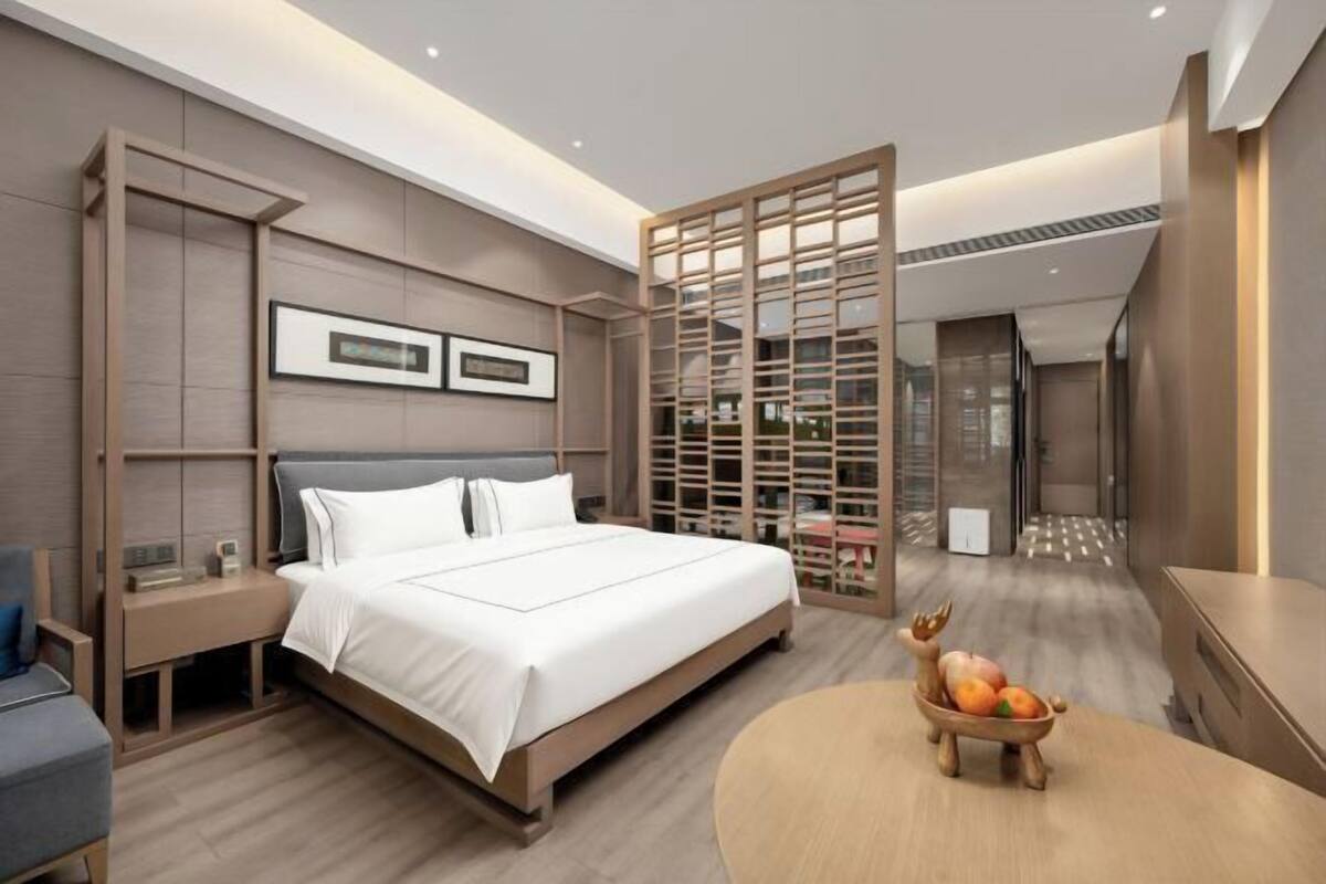 A modern hotel room with a large bed, bedside tables, a wooden partition, and a small round table with fruit.