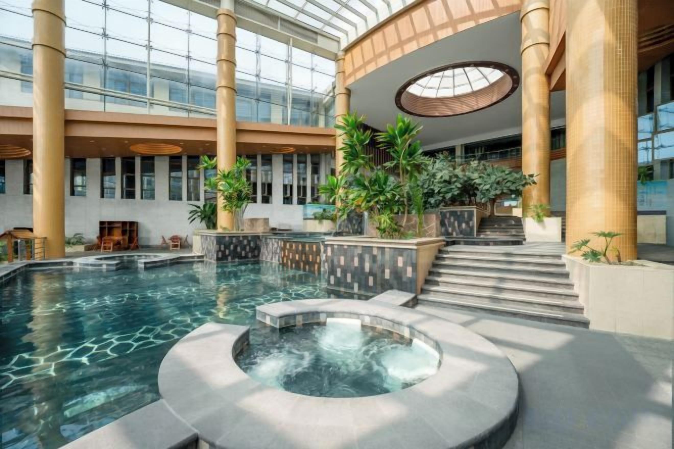 Una piscina interior con jacuzzi, rodeada de columnas y plantas, en un atrio amplio con techos altos y ventanas grandes.