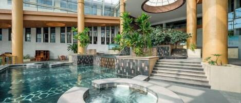 Una piscina interior con jacuzzi, rodeada de columnas y plantas, en un atrio amplio con techos altos y ventanas grandes.
