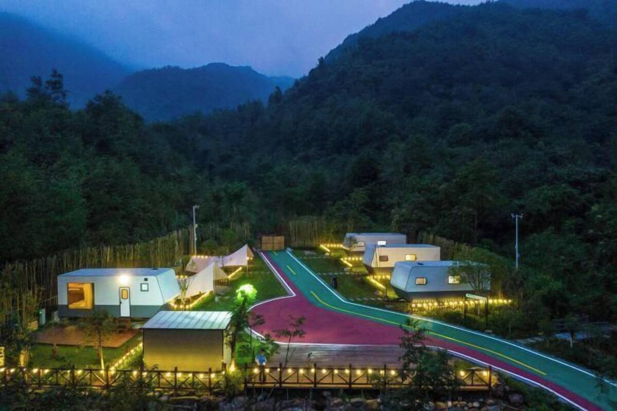 A series of modern, compact cabins with a paved road leading through a forested area.