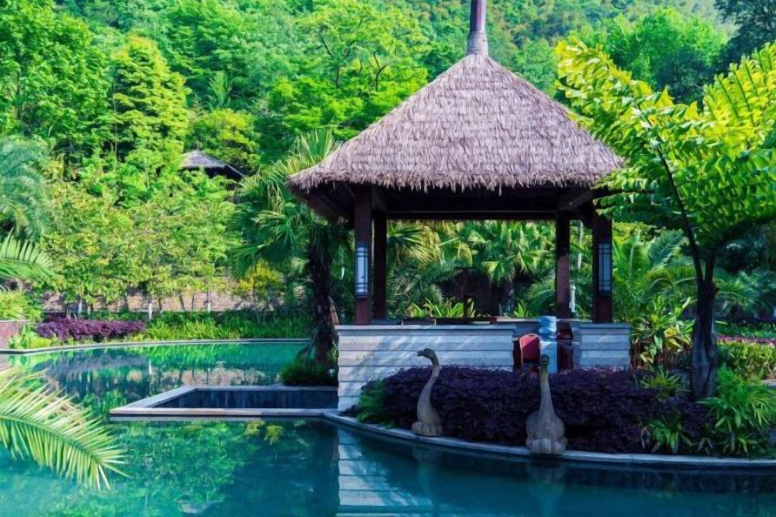 Un pabellón con techo de paja junto a una piscina, con un exuberante jardín tropical de fondo.
