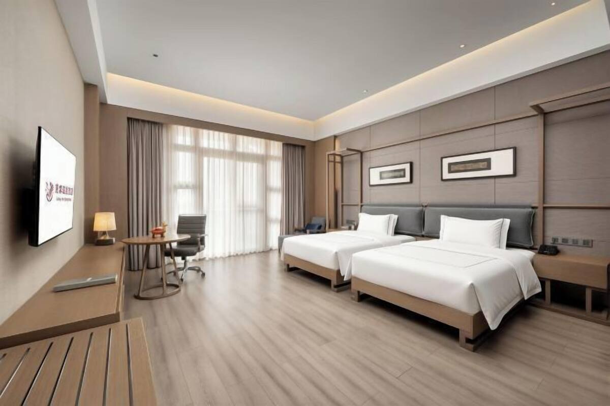 A hotel room with two beds, a desk, a chair, a TV, and a large window with curtains.