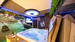 Een bubbelbad in een ruimte met een waterval op de achtergrond, een televisie en een houten tafel met kaarsen.