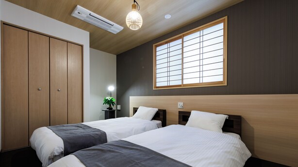 Een hotelkamer met twee bedden, een houten garderobe, een groot raam met zonwering en een plafondlamp.