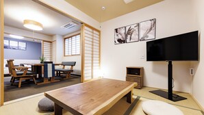 Een traditionele Japanse kamer met tatami vloeren, een lage houten tafel, een kussen van tatami stro en een televisie, met een schuifdeur naar een eetruimte.