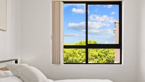 Een slaapkamer met een bed, een raam met uitzicht op groen en een gebouw, en een witte muur.