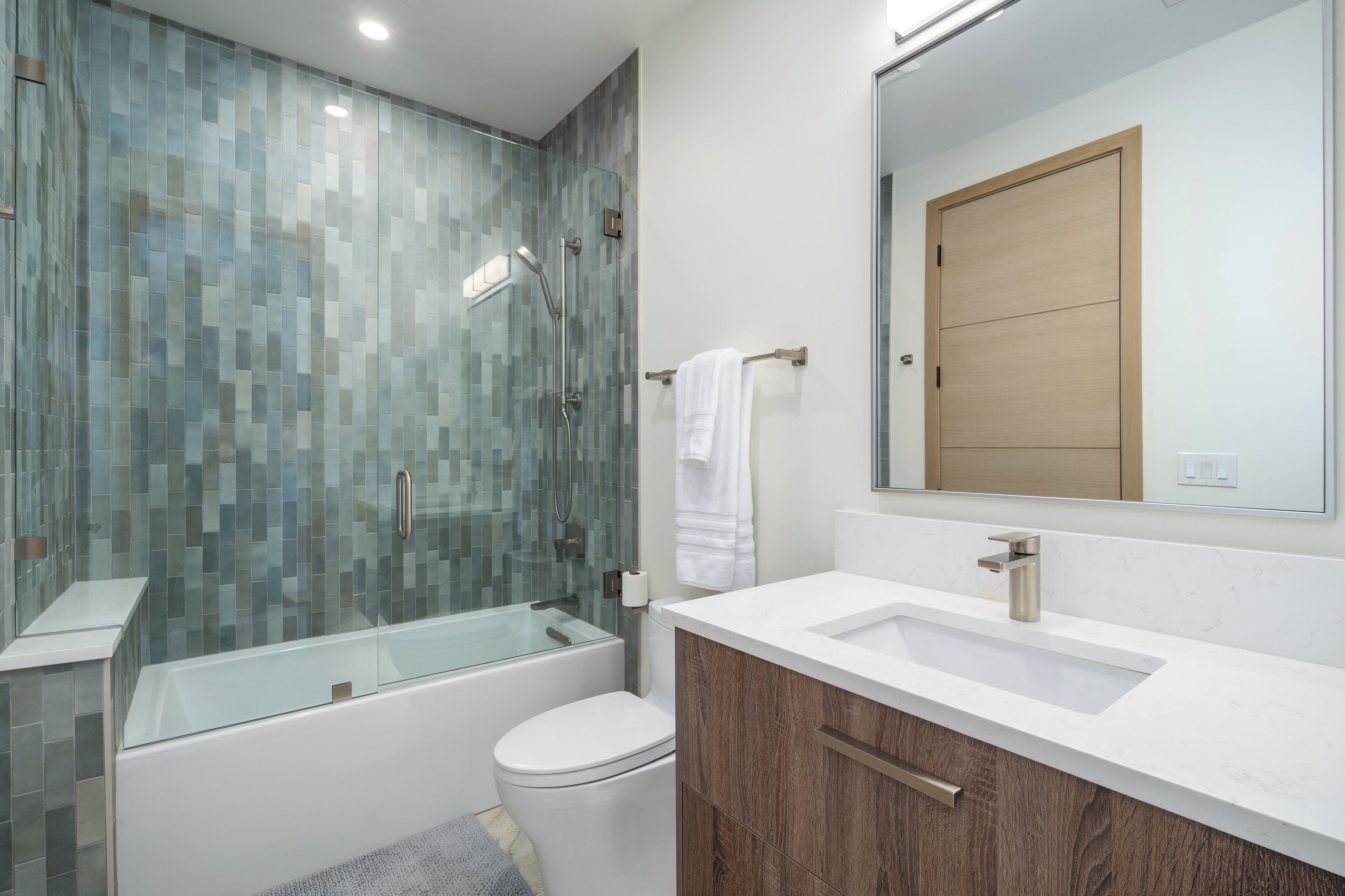 Un baño moderno con ducha envuelta en vidrio, inodoro blanco, tocador de madera con lavabo y espejo sobre el tocador.