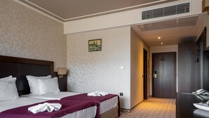 A hotel room with a large bed, a headboard, a bedside table, a lamp, a painting on the wall, and a door leading to another room.