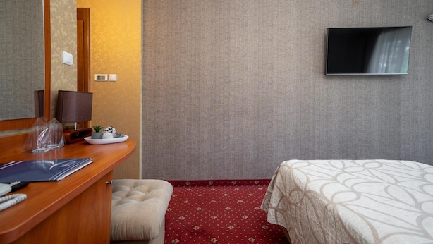 A hotel room with a wooden desk, a large mirror, a television on the wall, a bed with a patterned bedspread, and a red carpeted floor. - Spa Hotel Hissar (Hisarja)