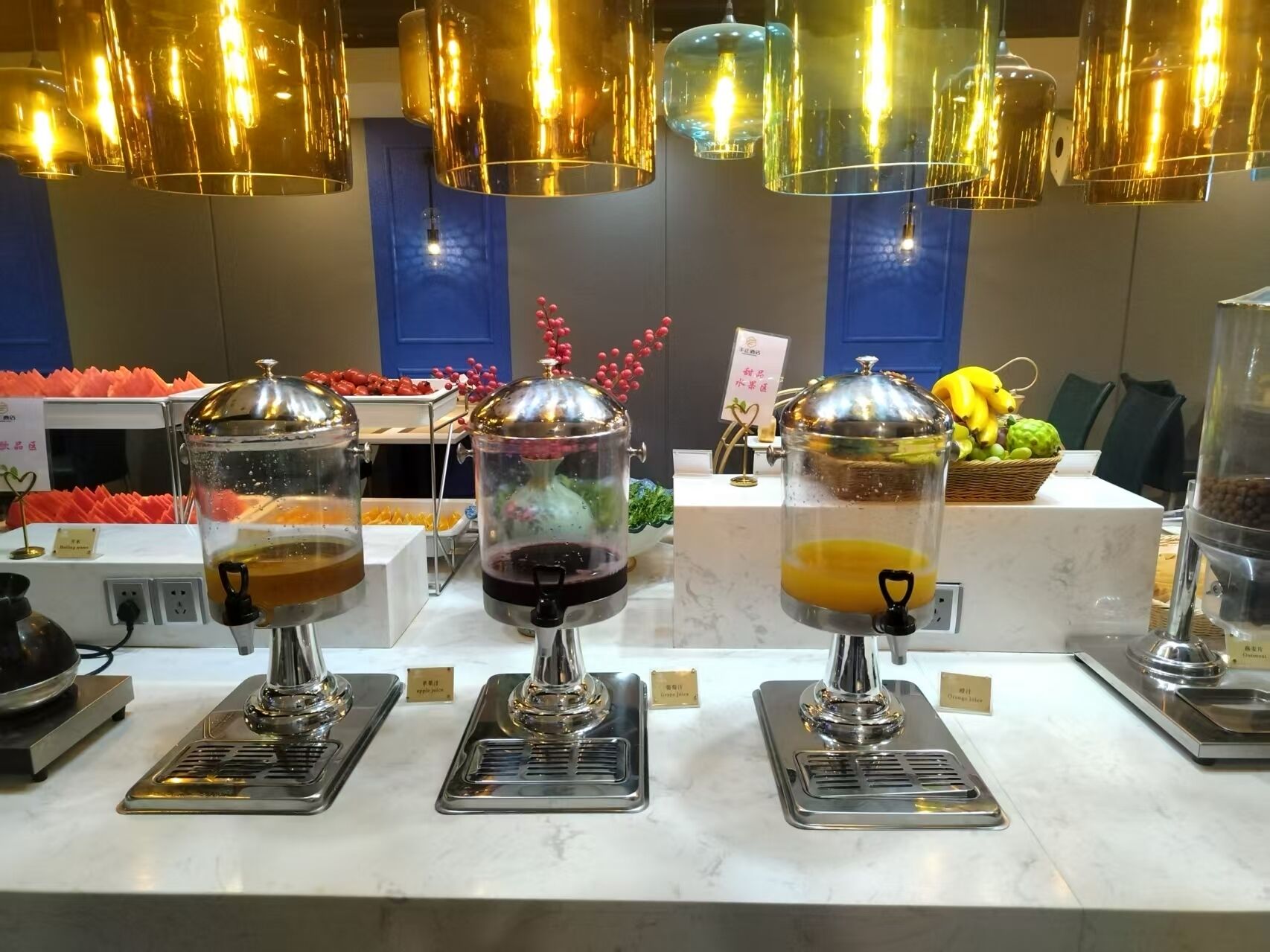 A buffet area with three beverage dispensers, a fruit basket, and a sign indicating the location.