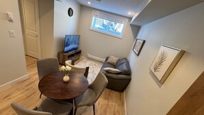 A modern living room with a dining table, grey chairs, a sofa, a TV, and a window with blinds. - Cozy  Nook (Saskatoon)
