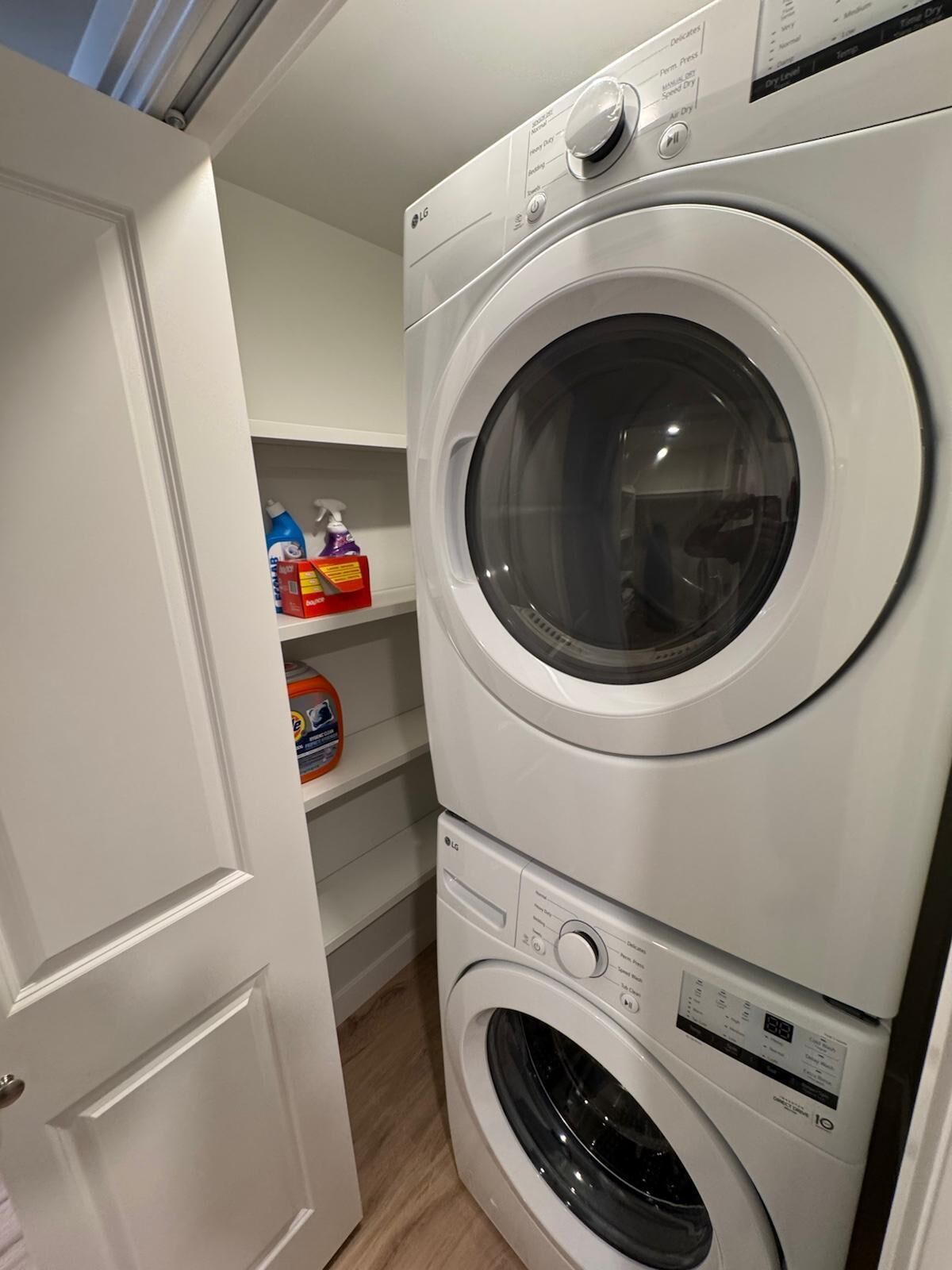 A laundry room with stacked washers and dryers, a shelf with cleaning supplies, and a white door.