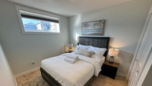 A bedroom with a bed, bedside lamps, a window, and a painting on the wall. - Cozy  Nook (Saskatoon)