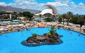 Ein großer Pool mit einer zentralen Insel, umgeben von Liegestühlen und Sonnenschirmen, mit einem markanten weißen Gebäude im Hintergrund.