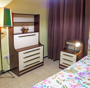 Ein Schlafzimmer mit einem Bett, einer Kommode mit Schubladen, einem Spiegel, einer Lampe und einem Fenster mit Vorhängen.