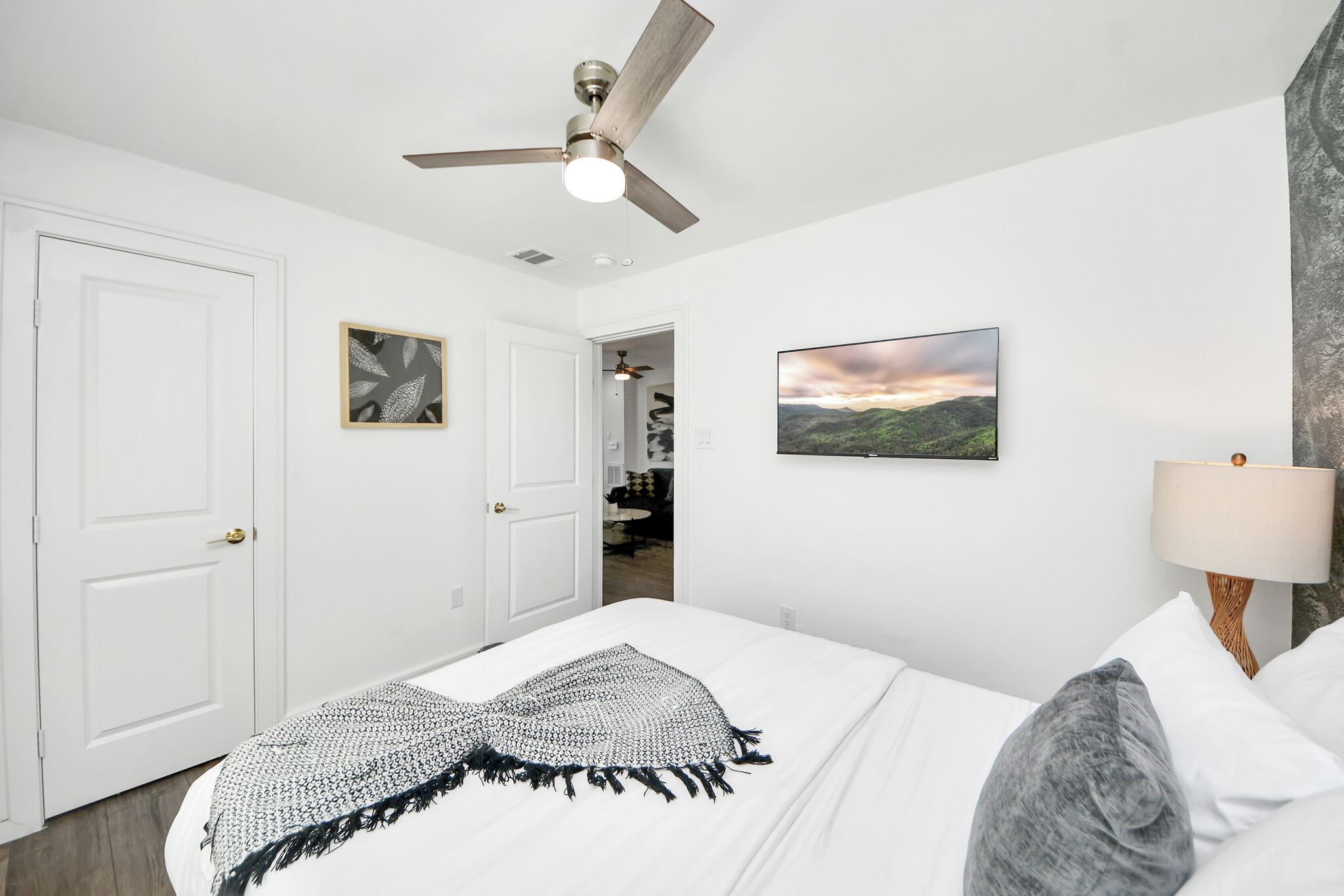 A bedroom with a bed, a ceiling fan, a nightstand with a lamp, a framed picture, and a door leading to a hallway.
