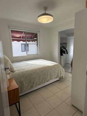 A bedroom with a bed, a window with curtains, a ceiling fan, and a door leading to another room.