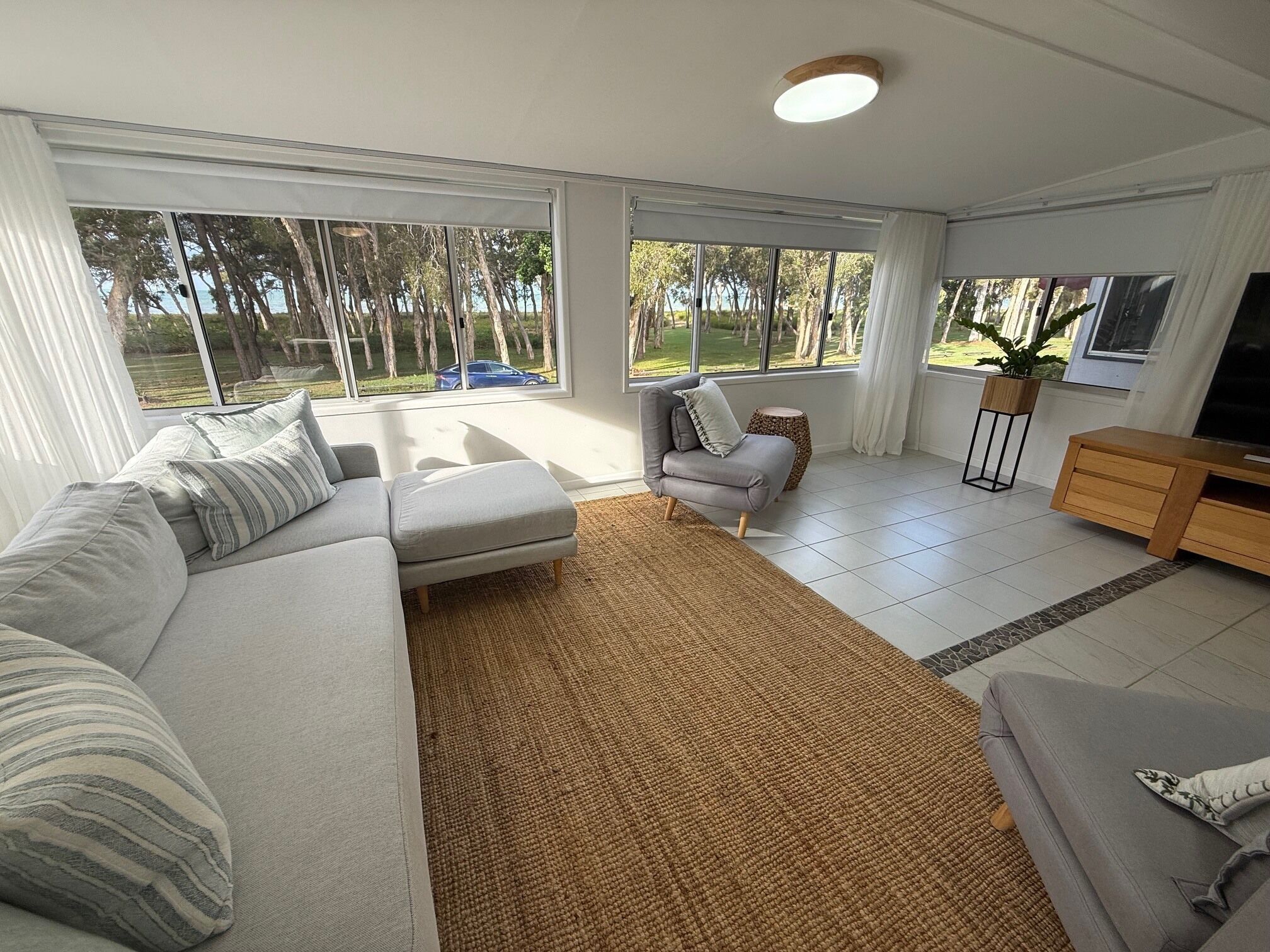 A modern living room with a sectional sofa, a small armchair, a TV stand, and large windows offering a view of the outdoors.