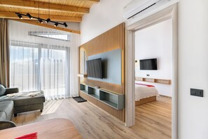 A modern hotel room with a wooden ceiling, a flat-screen TV mounted on a wooden panel, a sofa, a bed, and a large window with sheer curtains.