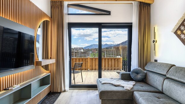 Een moderne woonkamer met een grote televisie, een bank en een schuifdeur naar een balkon met uitzicht op bergen.