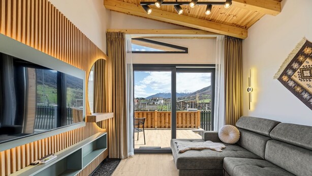 A modern living room with a large TV, a grey sofa, wooden paneling, and a balcony with a view of mountains.