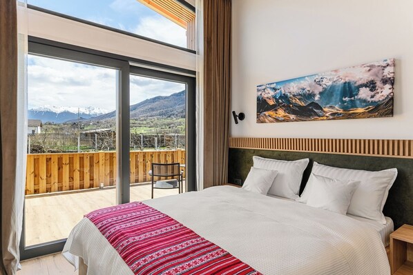 A bedroom with a large bed, a view of mountains, a wooden deck outside, and a painting on the wall.