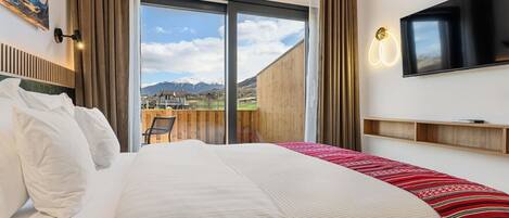 A hotel room with a large bed, a TV mounted on the wall, a view of mountains through a sliding door, and a wooden deck outside.