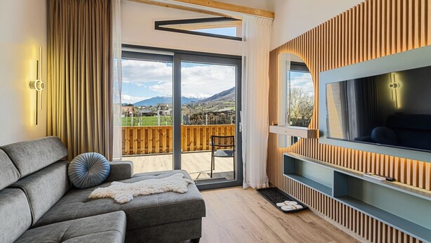 A modern living room with a grey sofa, a TV mounted on a wooden wall, and a sliding glass door leading to an outdoor patio. - Selva Cabin 1 - The Trailhead Retreat (Zărnești)