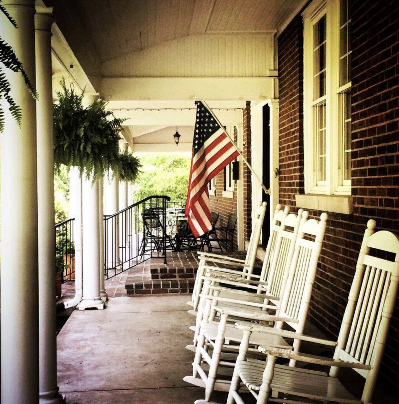 A porch with white rocking chairs, a brick building, and an American flag.