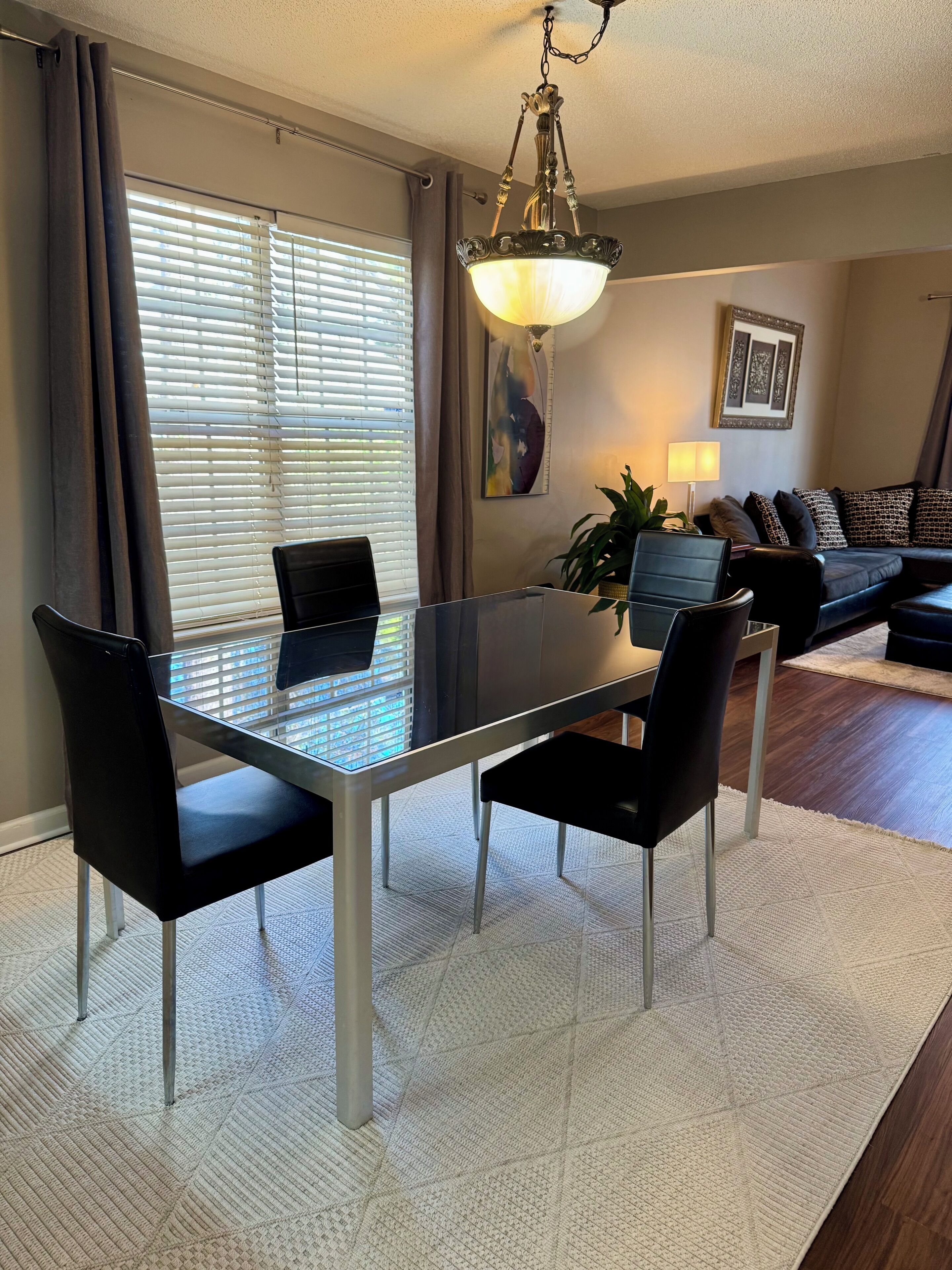 A dining area with a glass table, black chairs, a chandelier, a sofa, and a patterned rug.