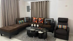 Ein Wohnzimmer mit einem Ecksofa, einem Glastisch, einem schwarzen Sessel und beigen Vorhängen.