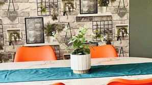 Un espace repas avec une table en marbre, des chaises orange et un mur décoré de plantes et d’un menu de cafés.