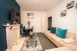 Een woonkamer met een bank, een aan de muur bevestigde televisie, een eettafel met stoelen en een deur naar een andere kamer.