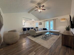 Un grand espace de vie comprenant un canapé, une table basse et des fauteuils, une cuisine à l’arrière-plan et un ventilateur de plafond.