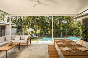 A modern outdoor living area with a pool, a sofa, a dining table set with plates and glasses, and a ceiling fan.