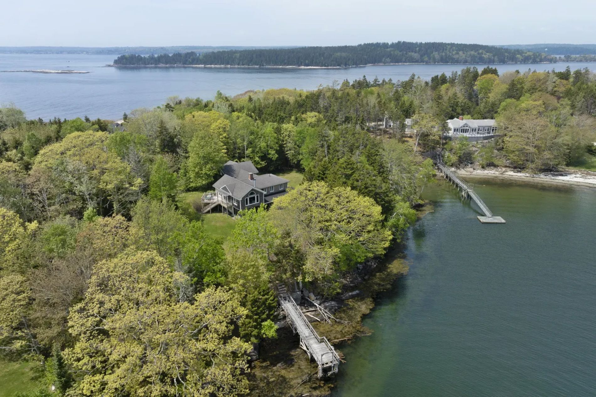 A large house surrounded by dense forest, a dock extending into a lake, and a distant shoreline.