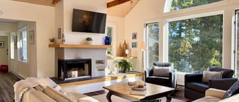 A spacious living room with a fireplace, comfortable seating, and large windows offering a view of the outdoors.