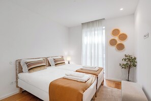 A hotel room with a bed, two bedside tables, a window with curtains, a wall-mounted light, and a small plant. - Sal D’Ouro Atlantic Living | Gym & Garage (Vila Nova de Gaia)