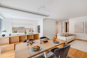 A modern kitchen with white cabinets, a dining area with a wooden table, and a living room with a sofa. - Sal D’Ouro Atlantic Living | Gym & Garage (Vila Nova de Gaia)