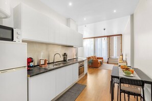 Eine moderne Küche mit weißen Schränken, einem eingebauten Backofen und einer Mikrowelle. Es gibt einen Essbereich mit Tisch und Stühlen, und ein Sofa im Hintergrund.