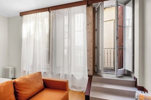 Ein Zimmer mit einer Couch, durchsichtigen Vorhängen, einer Heizung und Blick auf einen Balkon mit einem Tor.