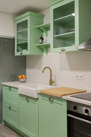 Een keuken met groene kastjes, een wit aanrecht, een houten snijplank, een roestvrijstalen spoelbak en een fornuis.