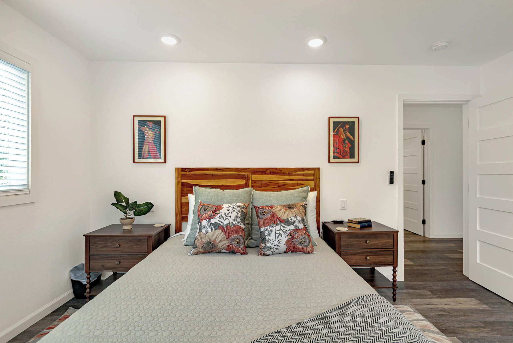 Une chambre à coucher comprenant un lit, deux tables de chevet, une tête de lit en bois, deux tableaux accrochés au mur, une plante en pot et une porte donnant sur une autre pièce.