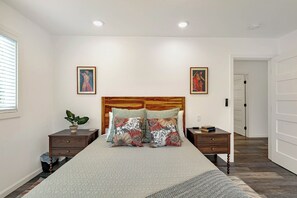 A bedroom with a bed, two nightstands, a wooden headboard, two wall-mounted paintings, a potted plant, and a door leading to another room.