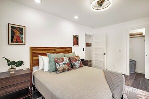 A bedroom with a wooden headboard, a bed with patterned pillows, a bedside plant, and a door leading to another room.