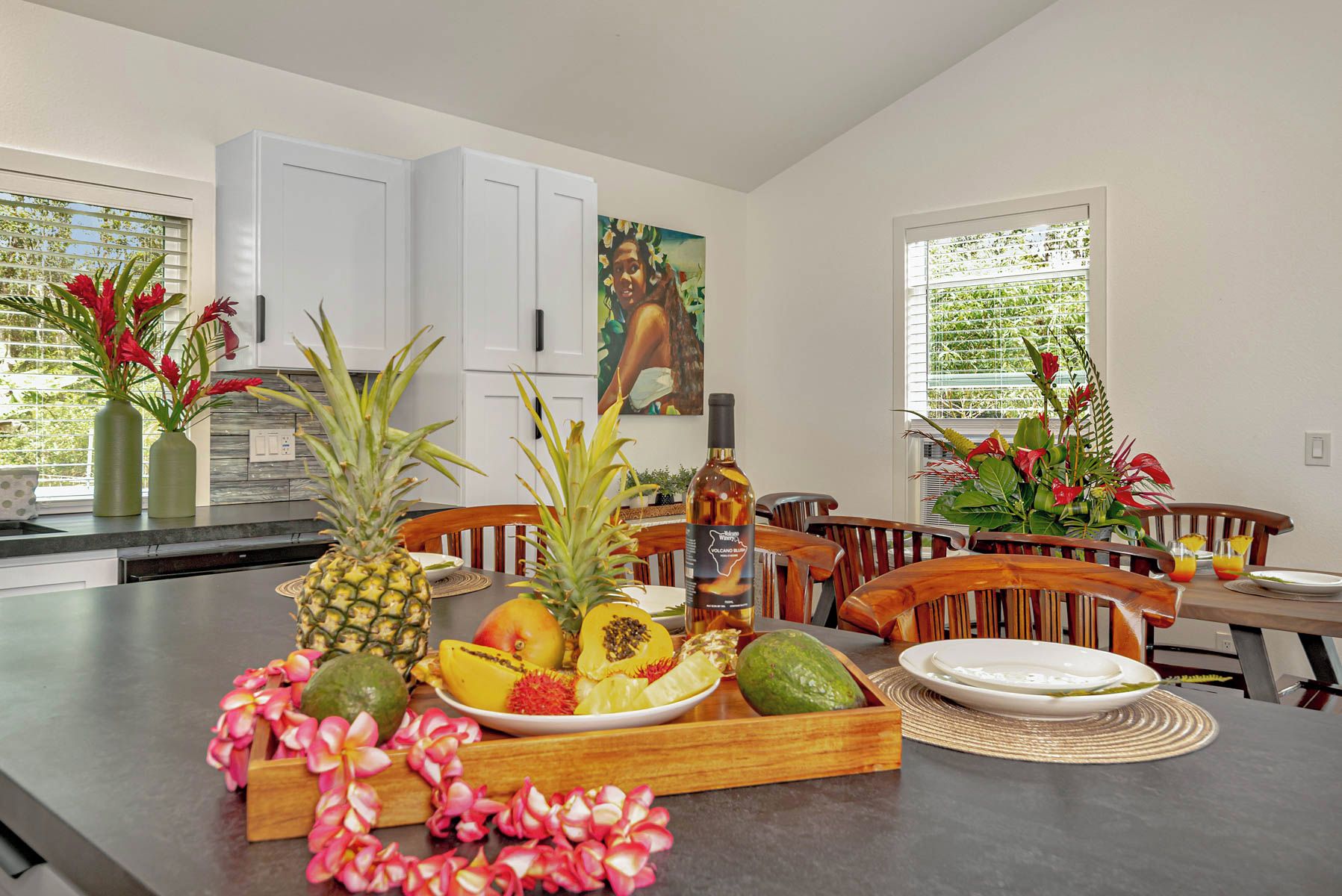 Une cuisine avec un présentoir pour les fruits et les bouteilles, des meubles blancs et un tableau accroché au mur.
